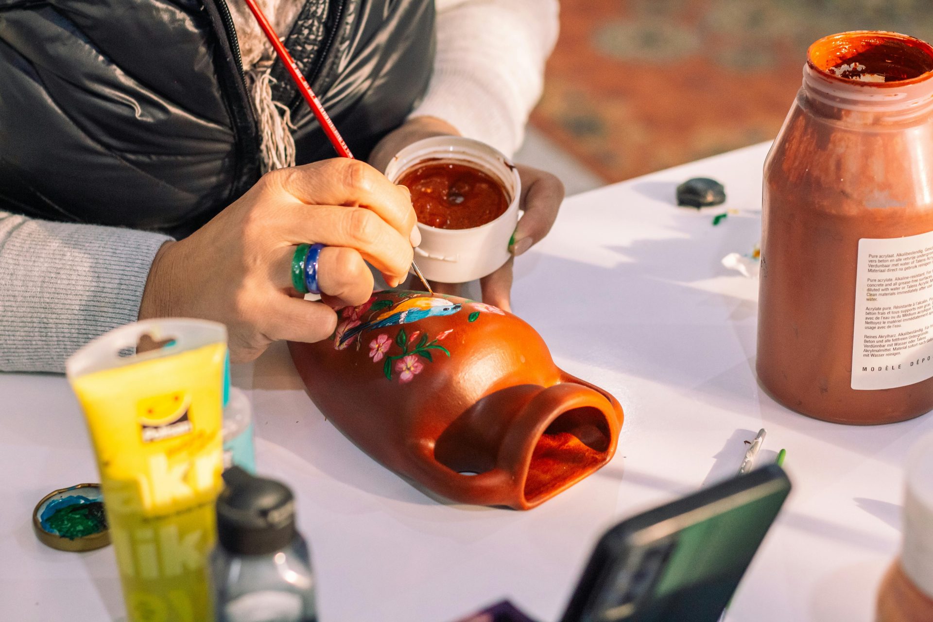 Artist hand painting a red vase with floral motifs using brushes and acrylic paint.